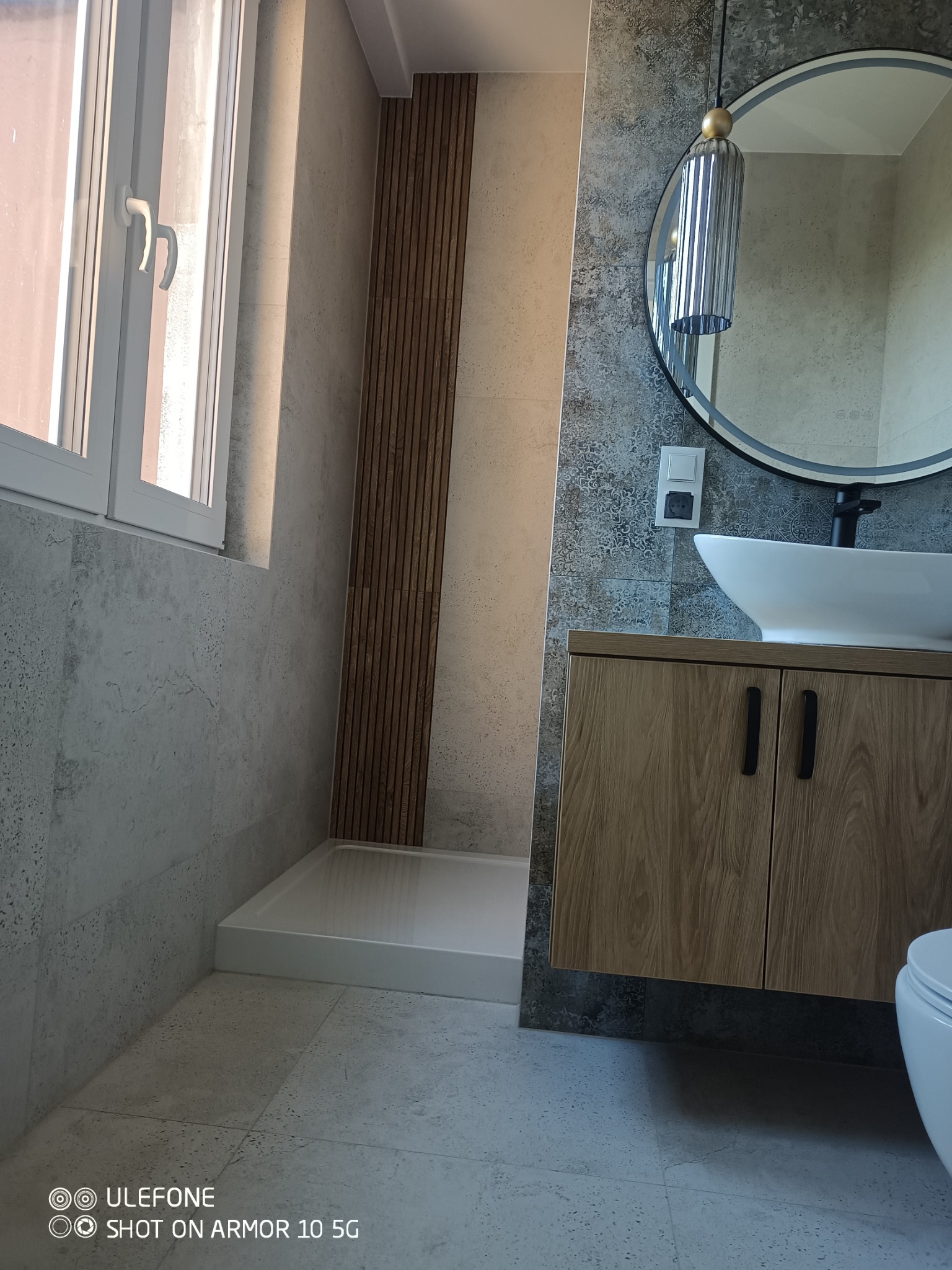 Nowoczesna łazienka po renowacji: szare płytki, drewniane akcenty, okrągłe lustro z lampą, biała umywalka nablatowa i kabina prysznicowa typu walk-in.