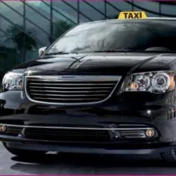 Czarny minivan z żółtym znakiem TAXI na dachu, zaparkowany na tle szklanego budynku, z numerem telefonu widocznym po lewej stronie.