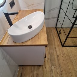 mark-bau - Nowoczesna łazienka z owalną, białą umywalką nablatową na blacie z drewnopodobnego materiału, czarna bateria, białe płytki imitujące marmur na ścianach i kabina prysznicowa z czarnymi profilami.