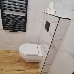 mark-bau - Nowoczesna łazienka z podwieszaną toaletą, grzejnikiem drabinkowym w kolorze czarnym i marmurowymi płytkami na ścianach oraz drewnopodobnymi płytkami na podłodze. Na gebericie ułożone trzy rolki...