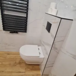 Nowoczesna łazienka z podwieszaną toaletą, grzejnikiem drabinkowym w kolorze czarnym i marmurowymi płytkami na ścianach oraz drewnopodobnymi płytkami na podłodze. Na gebericie ułożone trzy rolki...