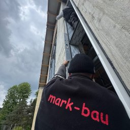 mark-bau - Montaż okna aluminiowego z użyciem poziomicy przez dwóch pracowników. Widoczny fragment elewacji i niebo. Na bluzie logo firmy 'mark-bau'.