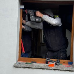 mark-bau - Montaż okna: pracownik wierci w drewnianej ramie okiennej, widoczne narzędzia na parapecie, drugi pracownik asystuje. Detal: okno z brązową ramą.