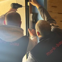 mark-bau - Montaż okna PCV z użyciem wkrętarki. Dwóch monterów w bluzach z logo firmy instaluje okno w niebieskim pomieszczeniu, widoczne listwy okienne Drutex.