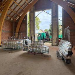 mark-bau - Paczki okien PCV w folii, przygotowane do montażu w surowym wnętrzu budynku z drewnianą konstrukcją dachu i widokiem na zewnątrz z zielonym autem.