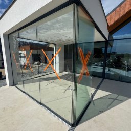 mark-bau - Nowoczesne, panoramiczne okna przesuwne z czarnymi ramami i pomarańczowymi oznaczeniami na szkle, widoczne z zewnątrz budynku. Minimalistyczna architektura.