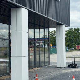 mark-bau - Nowoczesny budynek z przeszklonym wejściem i kolumnami obłożonymi białymi panelami. Widoczne elementy wykończeniowe i pomarańczowe pachołki.