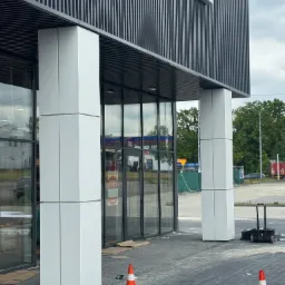 Nowoczesny budynek z przeszklonym wejściem i kolumnami obłożonymi białymi panelami. Widoczne elementy wykończeniowe i pomarańczowe pachołki.