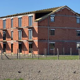 mark-bau - Surowy, trzypiętrowy budynek z cegły z zamontowanymi oknami i drewnianą konstrukcją dachu w trakcie budowy, ogrodzony drewnianym płotem na tle błękitnego nieba.