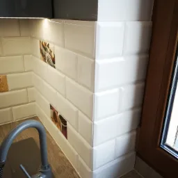 Białe, błyszczące płytki ułożone w cegiełkę, z dekoracyjnymi wstawkami z motywami jedzenia, szara bateria kuchenna i fragment okna z brązową ramą, na blacie imitującym drewno.