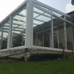 Szklana, biała altana przylegająca do ceglanego domu z widocznym orynnowaniem i zielonym trawnikiem.