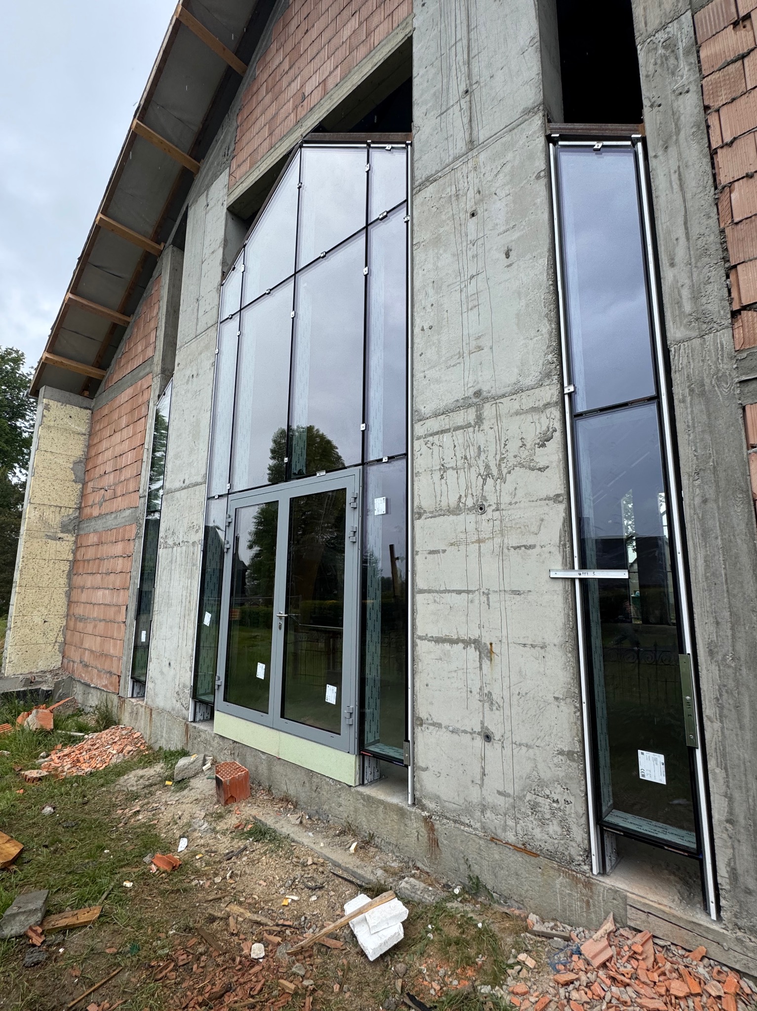 Montaż okien aluminiowych w surowym budynku. Widoczne duże przeszklenia, cegły i beton. Częściowo zamontowane okna w szarej ramie. Teren budowy, fragment trawy.