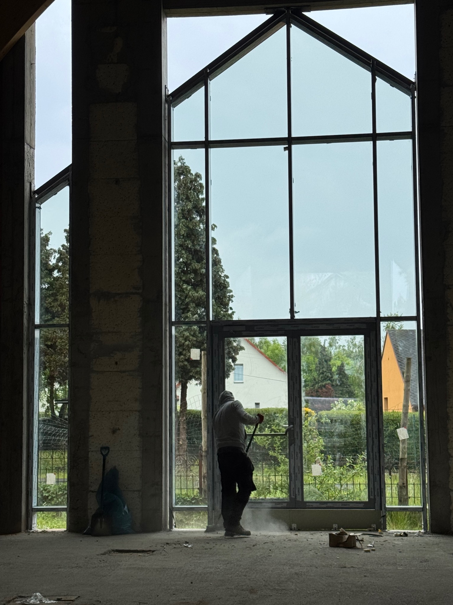 Montaż okien trapezowych w surowym stanie budynku. Widoczne narzędzia i osoba montująca okno. Duże przeszklenia z widokiem na zieleń.