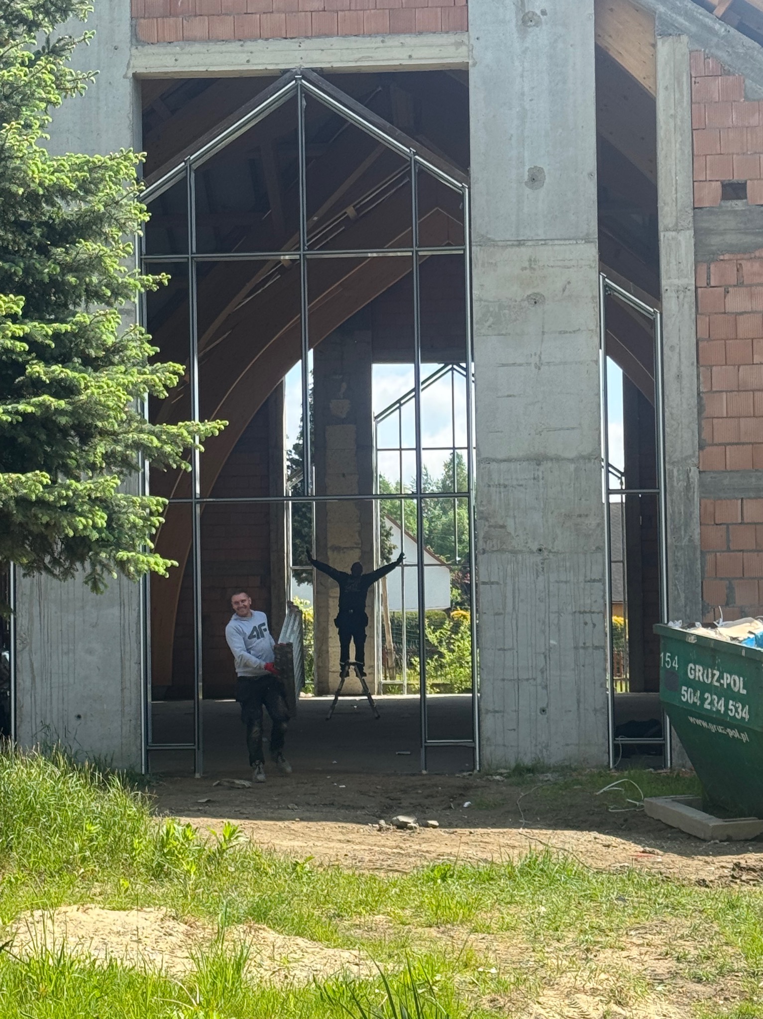 Montaż konstrukcji okiennej z metalu w surowym budynku. Dwóch mężczyzn przy pracy, jeden na drabinie. Widoczny kontener na gruz i drzewo iglaste.