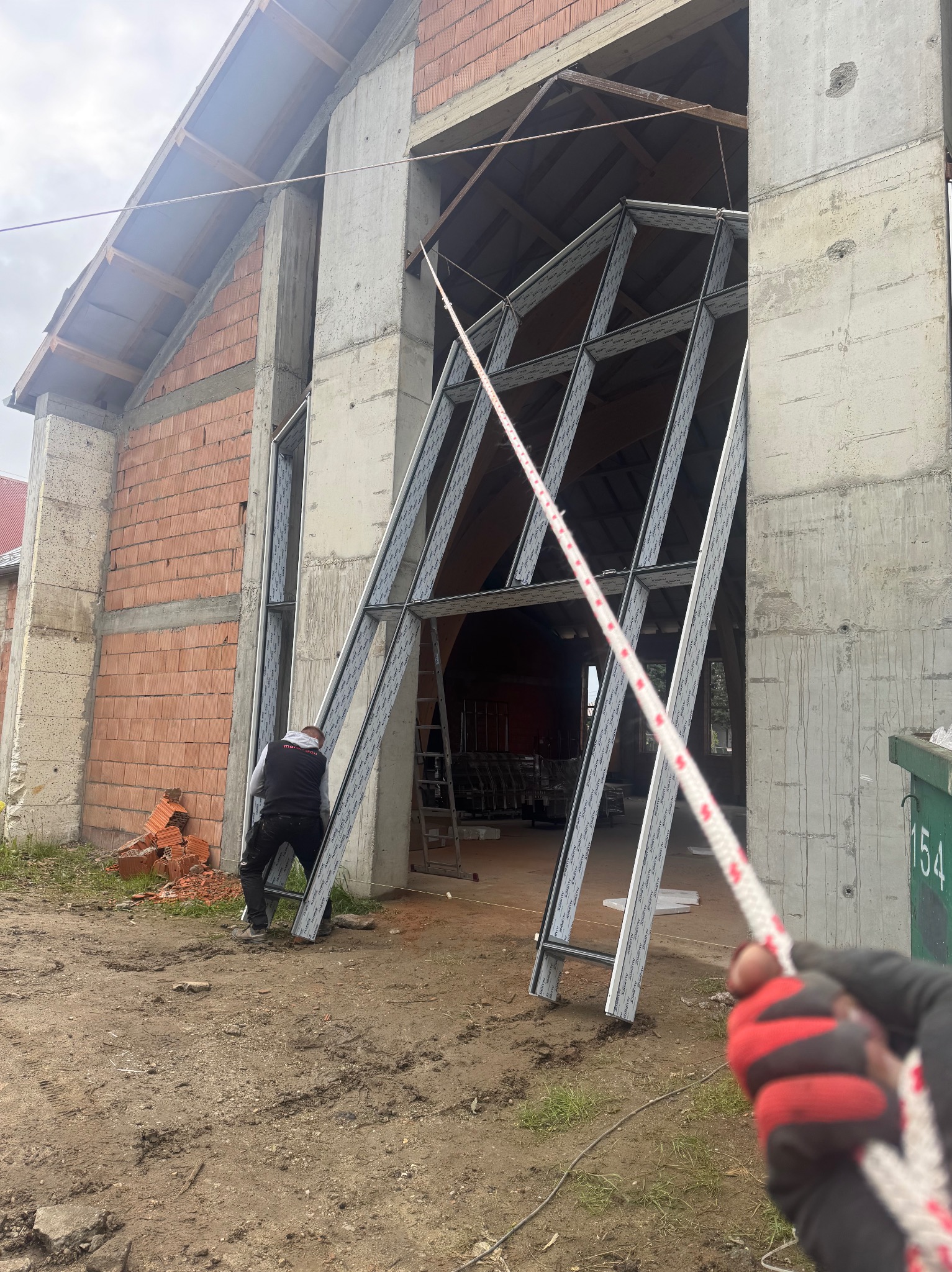 Montaż konstrukcji okiennej w surowym budynku. Widoczny pracownik, lina i fragment dłoni w rękawicy. Budynek w stanie deweloperskim z cegły i betonu.