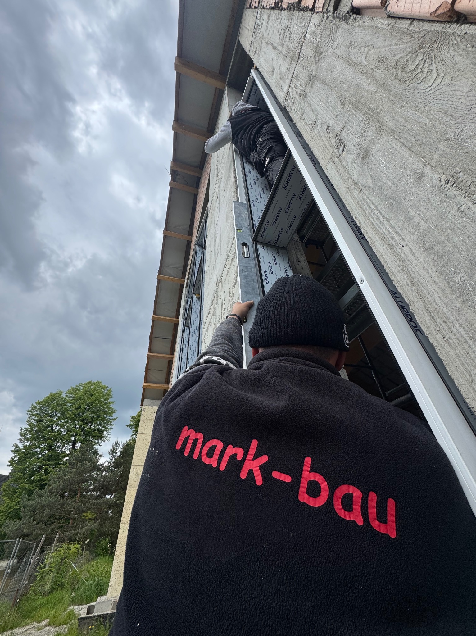 Montaż okna aluminiowego z użyciem poziomicy przez dwóch pracowników. Widoczny fragment elewacji i niebo. Na bluzie logo firmy 'mark-bau'.
