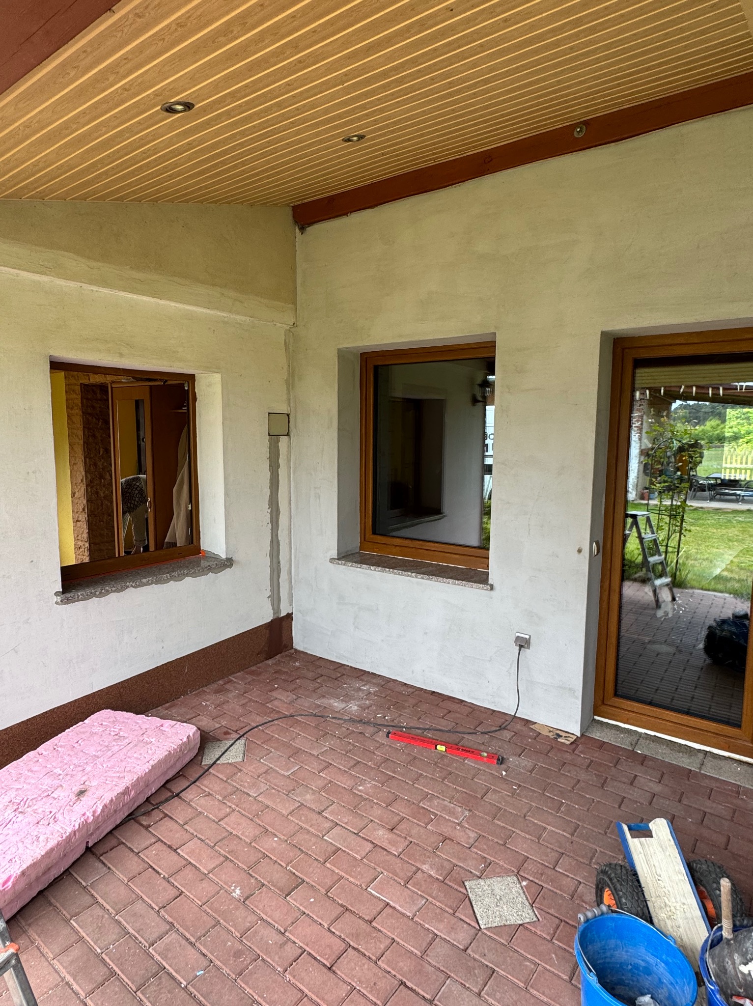 Montaż okien z brązowymi ramami w surowym wnętrzu. Widoczne narzędzia pracy, ceglana podłoga i drewniana podsufitka. Etap prac wykończeniowych.