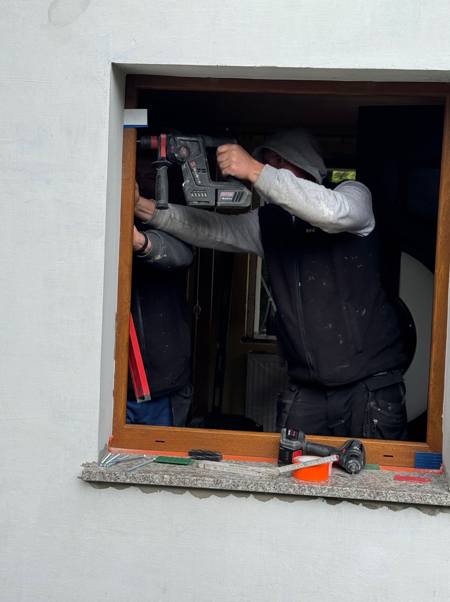 Montaż okna: pracownik wierci w drewnianej ramie okiennej, widoczne narzędzia na parapecie, drugi pracownik asystuje. Detal: okno z brązową ramą.