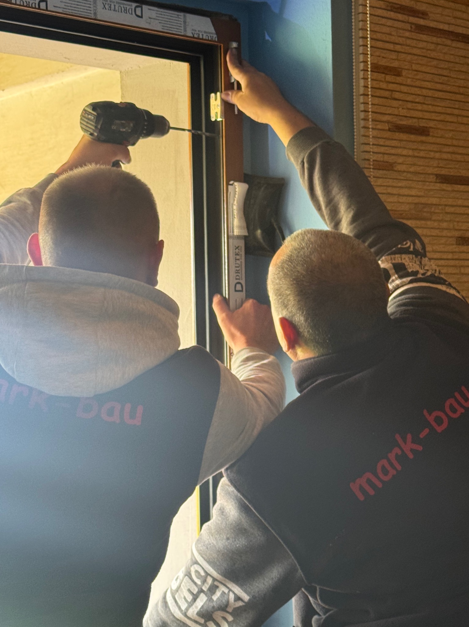 Montaż okna PCV z użyciem wkrętarki. Dwóch monterów w bluzach z logo firmy instaluje okno w niebieskim pomieszczeniu, widoczne listwy okienne Drutex.