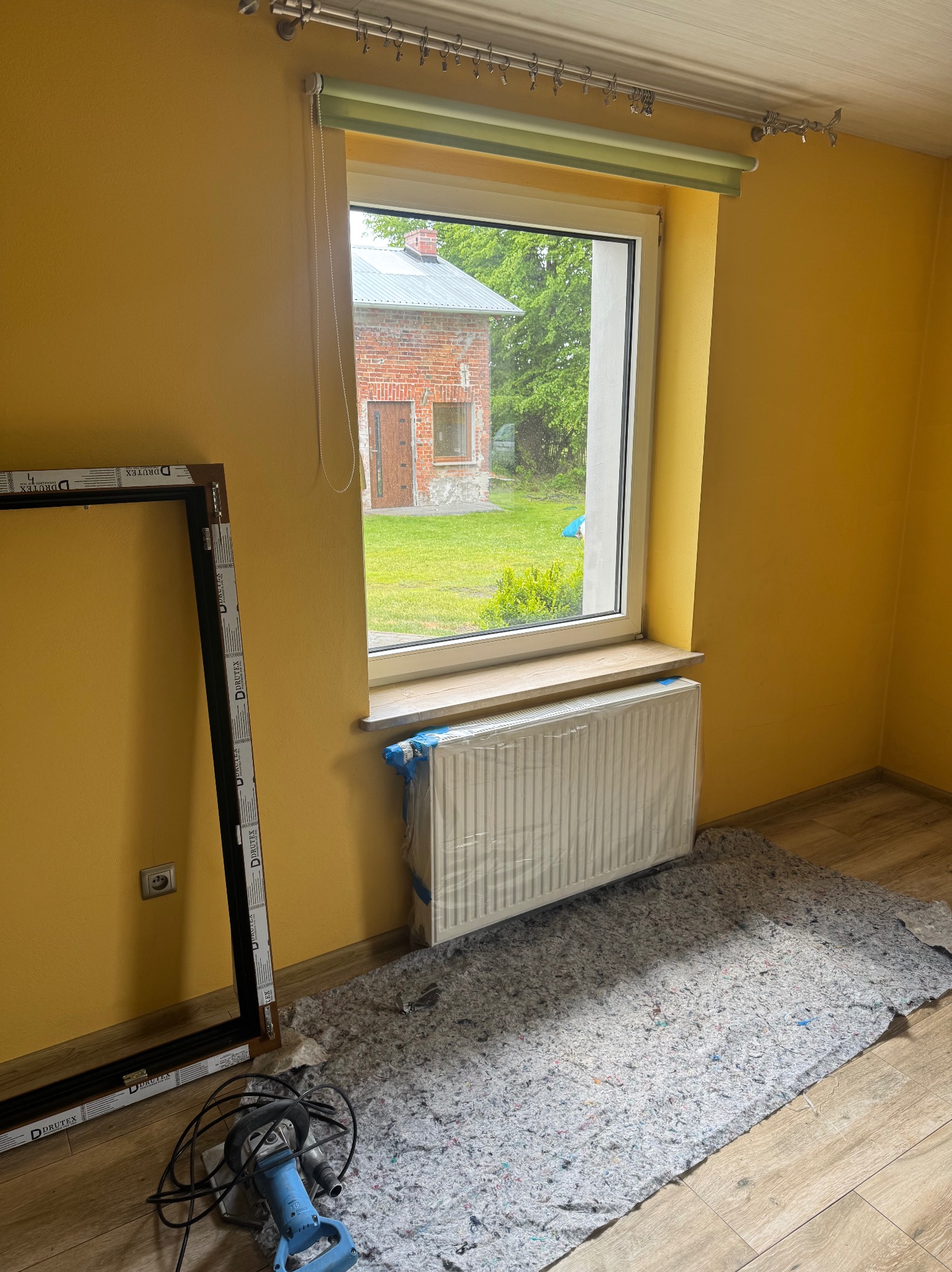 Żółty pokój w trakcie wymiany okna PCV. Nowa rama okienna oparta o ścianę, grzejnik zabezpieczony folią, widoczne narzędzia. Na zewnątrz budynek z czerwonej cegły.