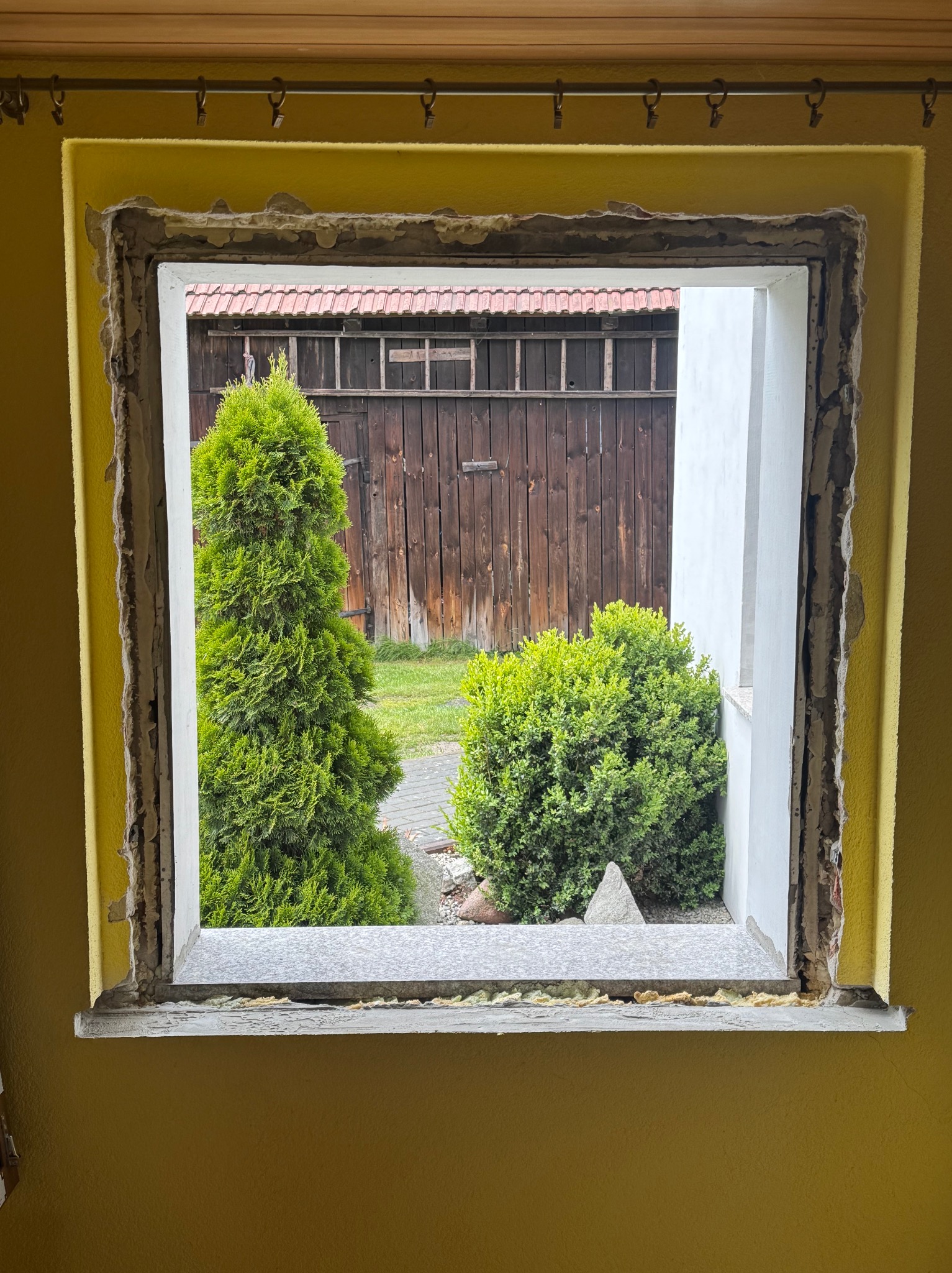 Otwór okienny w trakcie montażu, widoczne uszczelnienia i surowa ściana. Na zewnątrz zieleń: tuja i krzew, płot z desek. Ściana w odcieniach żółci, widoczne ślady po starym oknie.
