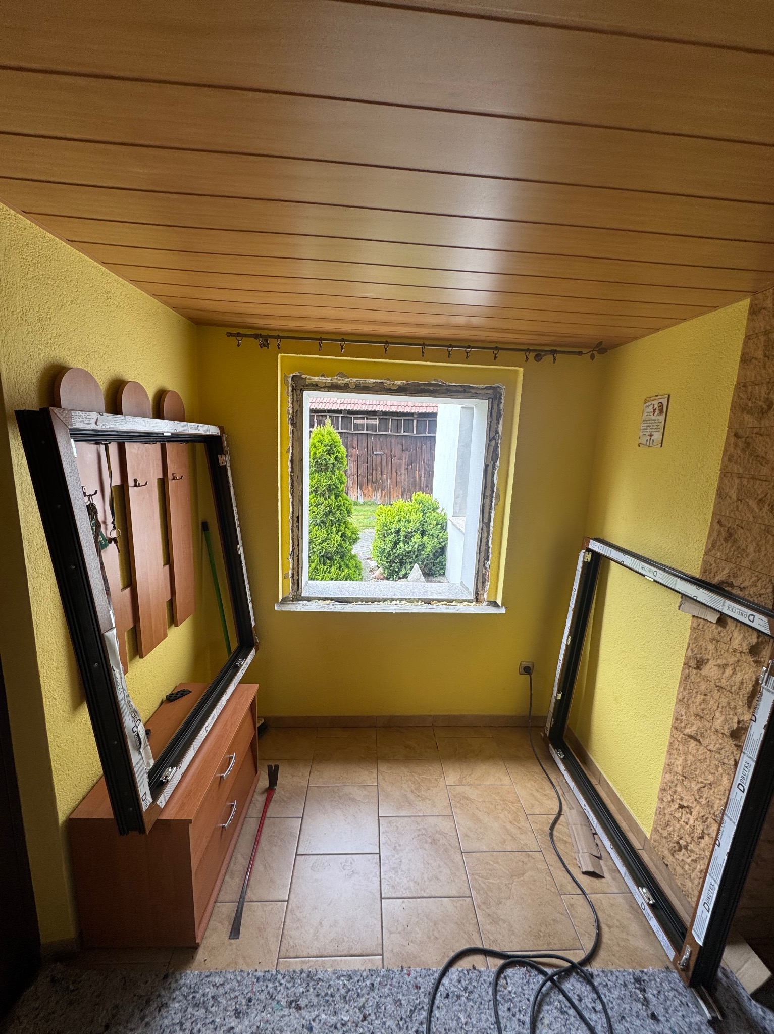 Żółte wnętrze pokoju z widokiem na zieleń przez okno bez ramy. Dwie ramy okienne oparte o ściany. Widoczny etap wymiany okna w domu.