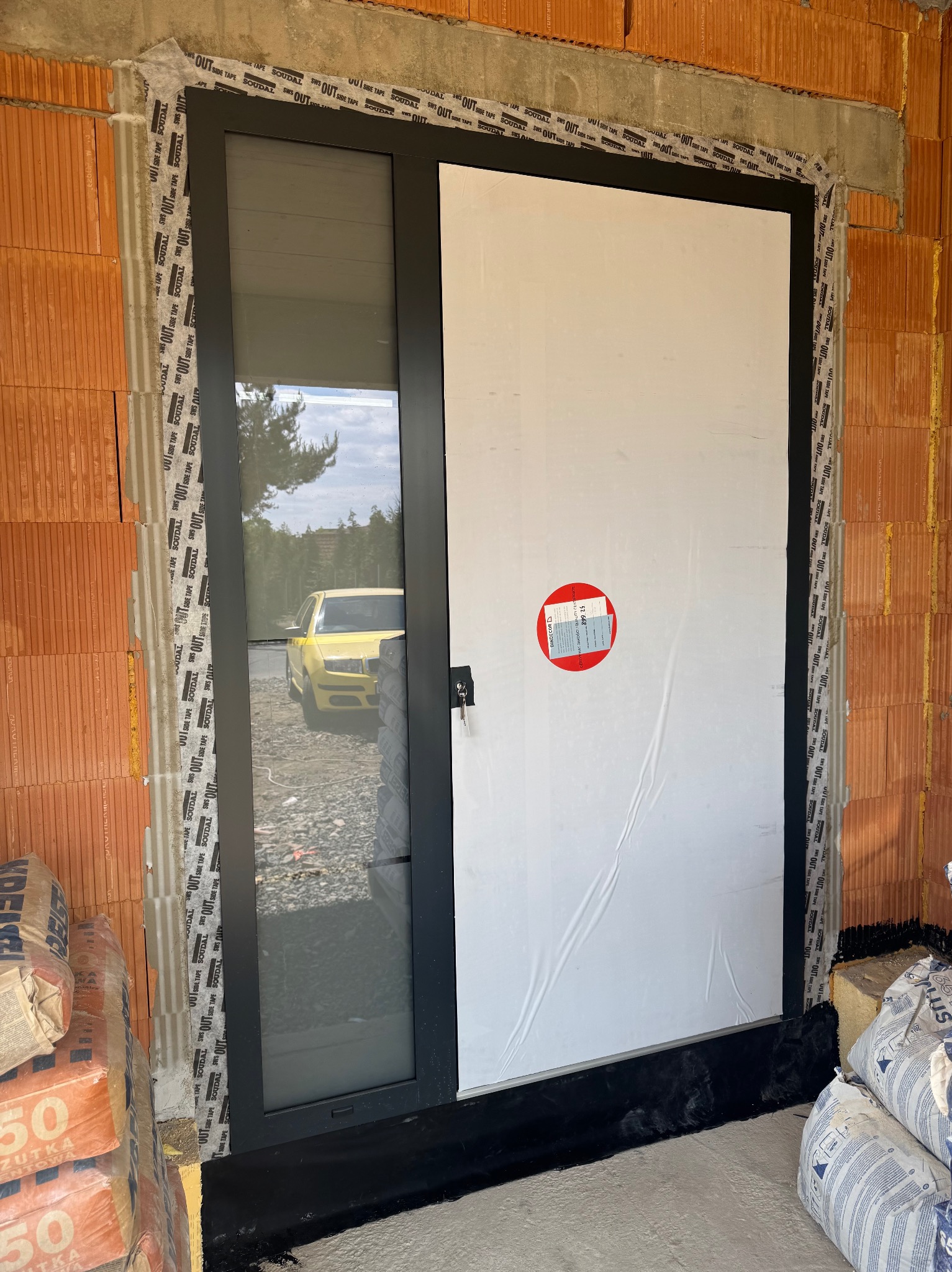 Montaż okna z czarną ramą w surowym murze z cegły, zabezpieczonego taśmą izolacyjną. Widoczne worki z zaprawą murarską i odbicie żółtego samochodu w szybie.