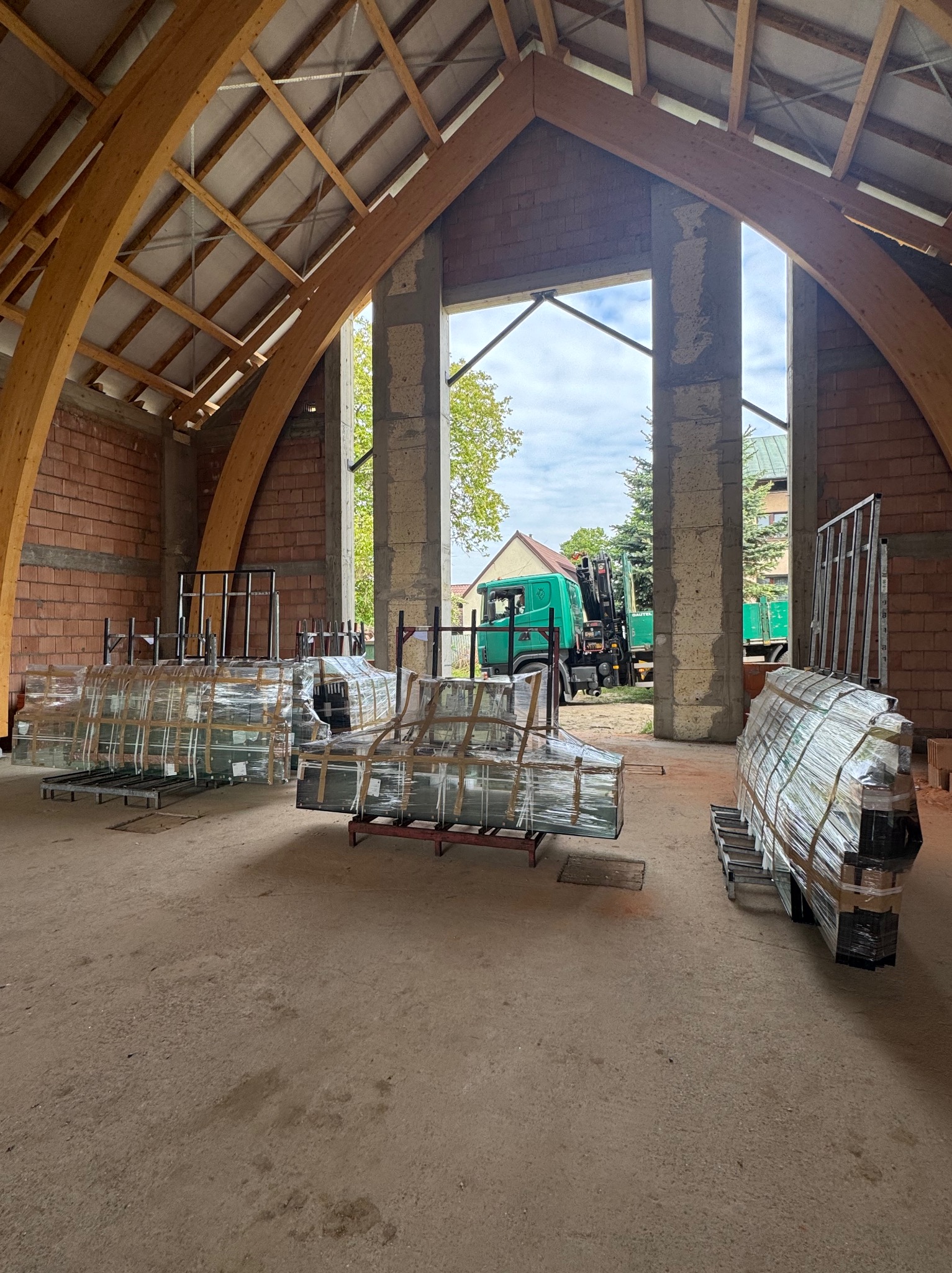 Paczki okien PCV w folii, przygotowane do montażu w surowym wnętrzu budynku z drewnianą konstrukcją dachu i widokiem na zewnątrz z zielonym autem.