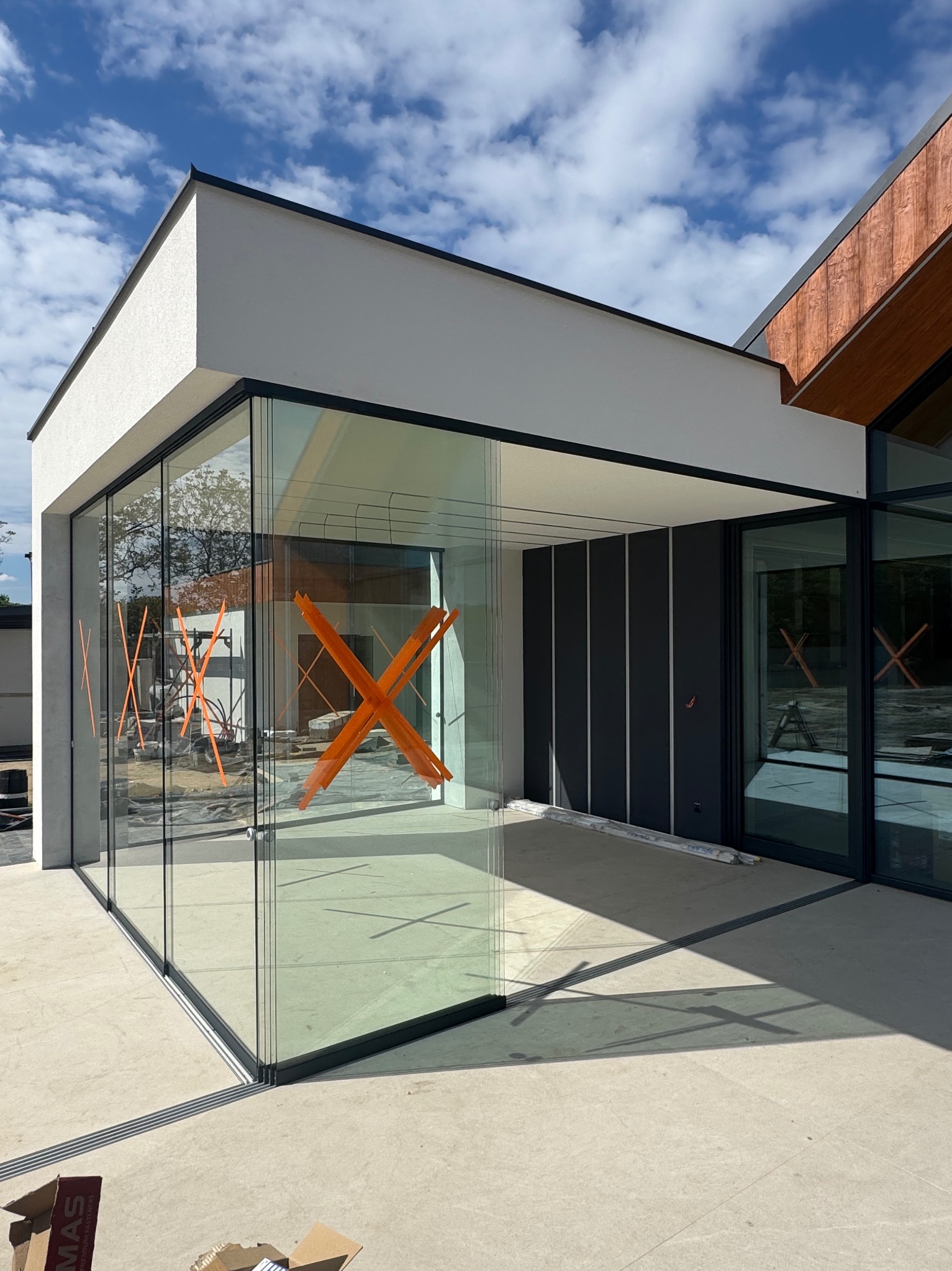 Nowoczesne, przeszklone narożne okno z czarnymi ramami, zabezpieczone pomarańczową taśmą krzyżową, w jasnym budynku z elementami drewnianymi. Błękitne niebo w tle.