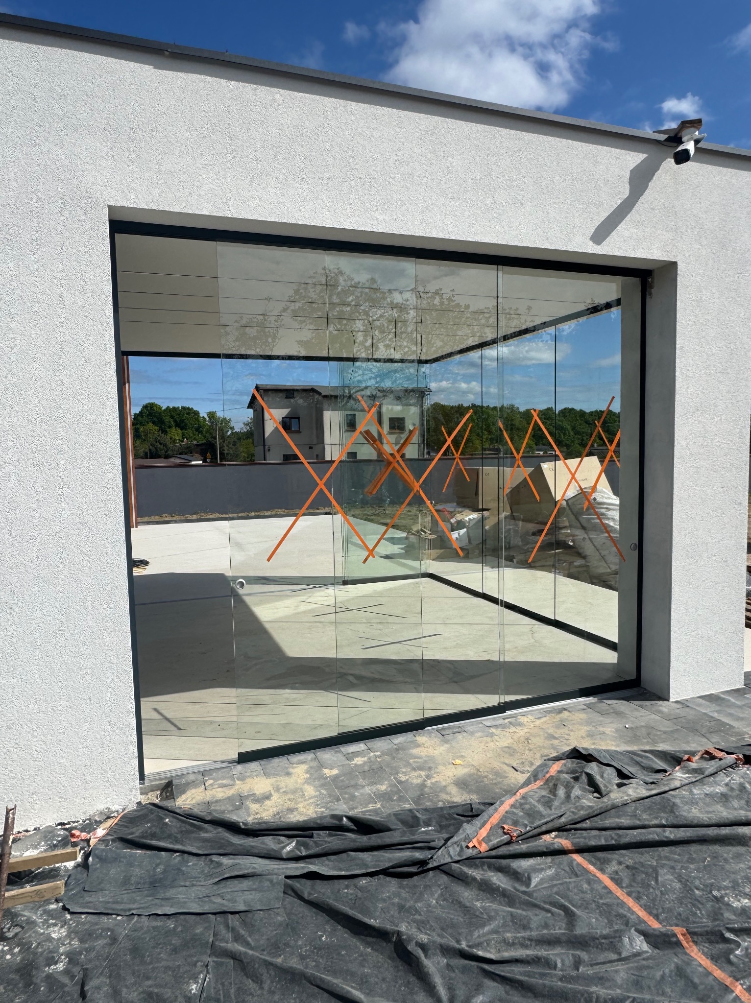 Nowoczesne, szklane drzwi przesuwne z czarną ramą w jasnym budynku. Pomarańczowe taśmy zabezpieczające na szybach. Widoczna instalacja kamery na ścianie.