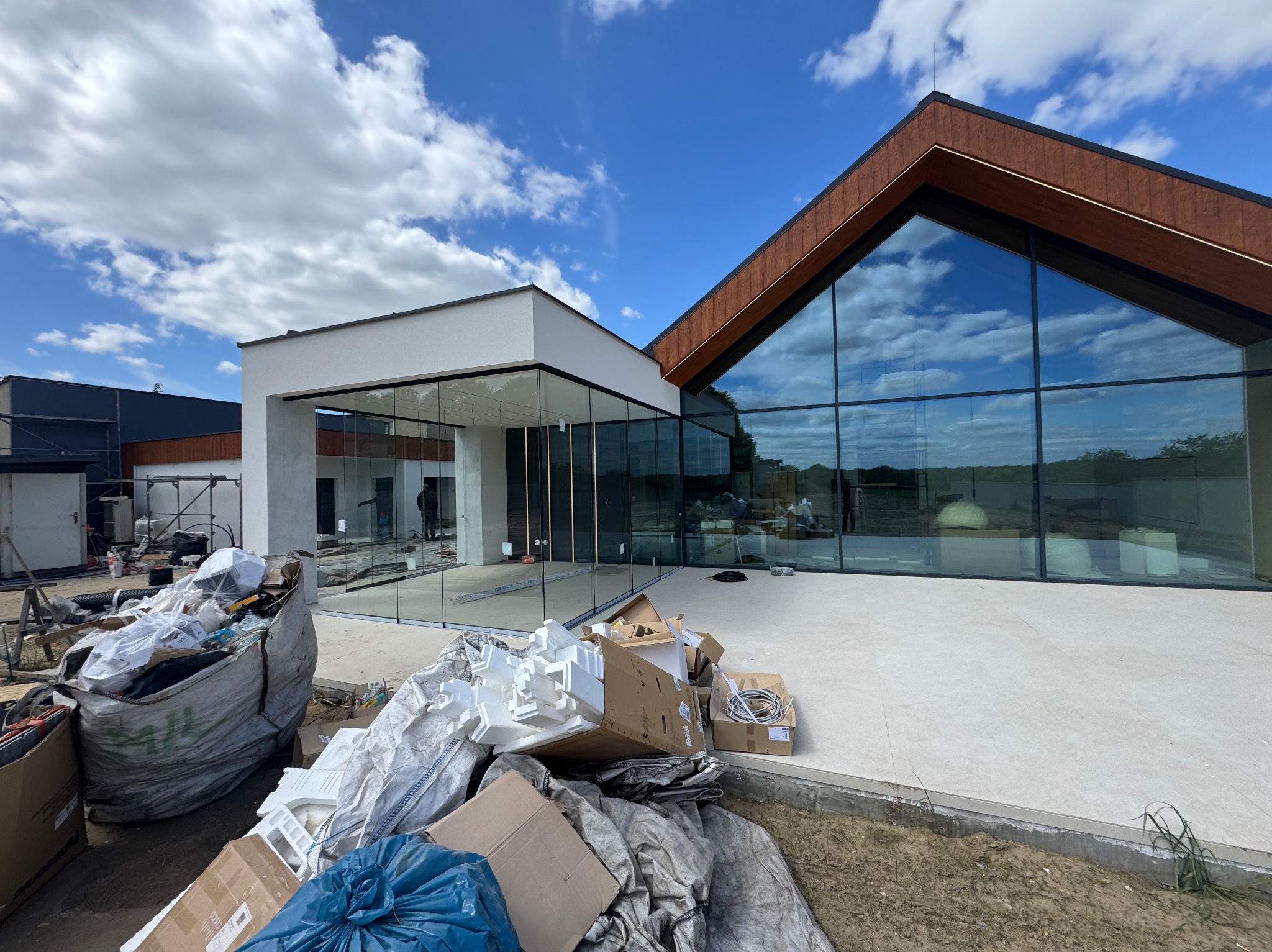 Nowoczesny budynek z dużymi przeszkleniami, w trakcie wykończenia. Widoczne materiały budowlane i odpady po montażu okien. Błękitne niebo z chmurami w odbiciu szyb.