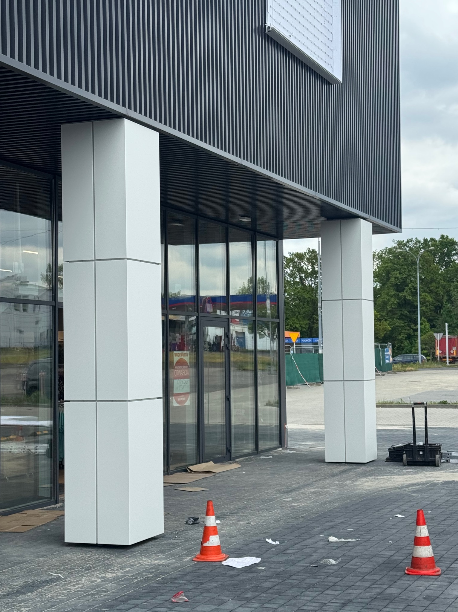 Nowoczesny budynek z przeszklonym wejściem i kolumnami obłożonymi białymi panelami. Widoczne elementy wykończeniowe i pomarańczowe pachołki.