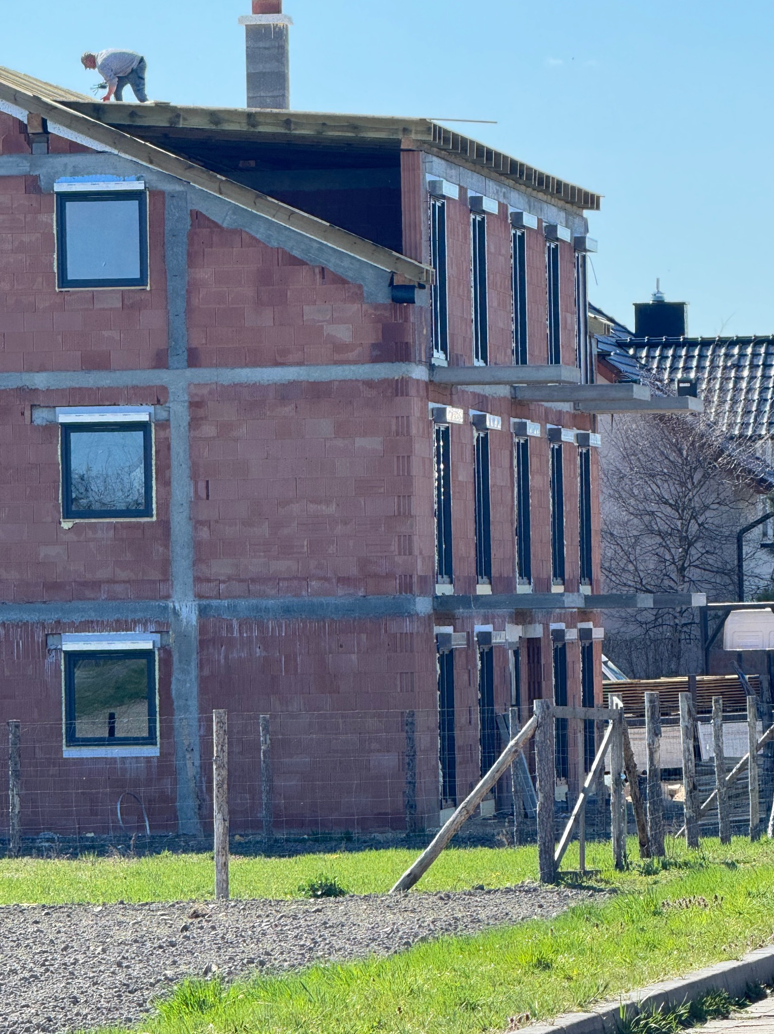 Budowa domu z czerwonej cegły z zamontowanymi oknami, widok na plac budowy, gdzie robotnik pracuje na dachu. W tle inne budynki.