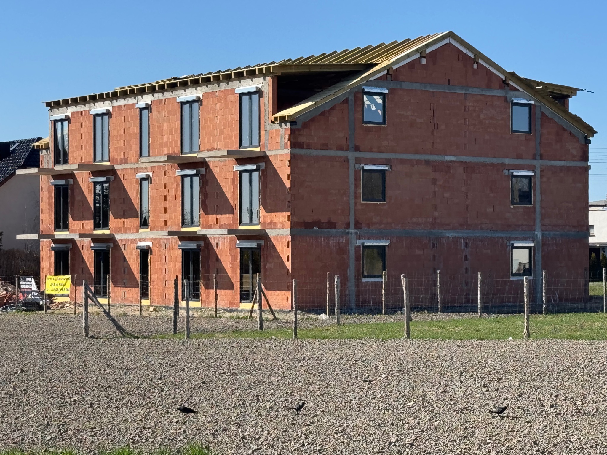 Surowy, trzypiętrowy budynek z cegły z zamontowanymi oknami i drewnianą konstrukcją dachu w trakcie budowy, ogrodzony drewnianym płotem na tle błękitnego nieba.