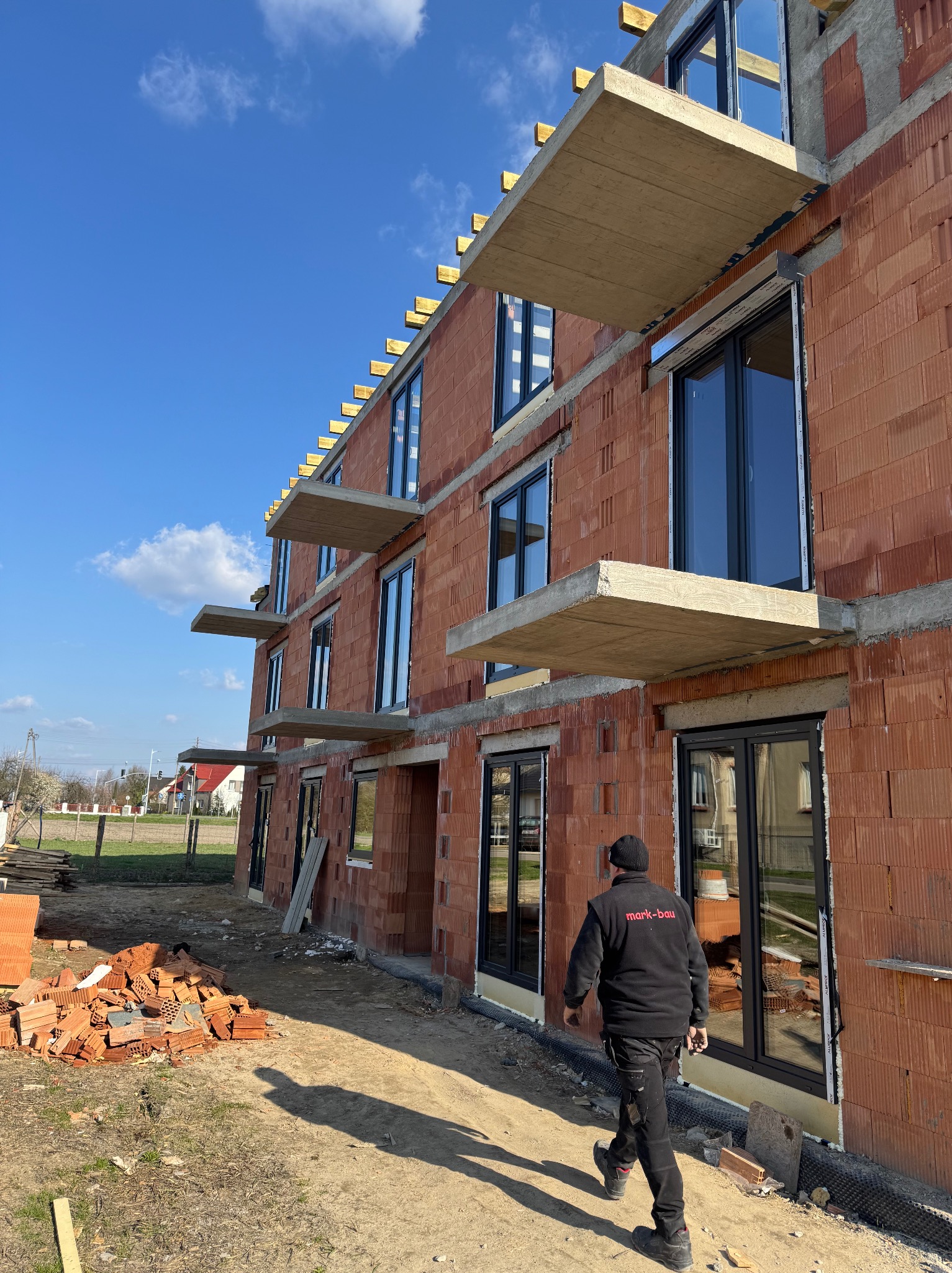 Budowa budynku z czerwonej cegły ceramicznej z zamontowanymi oknami PCV, widoczne betonowe nadproża, pracownik w czarnej kurtce i sterta cegieł obok.