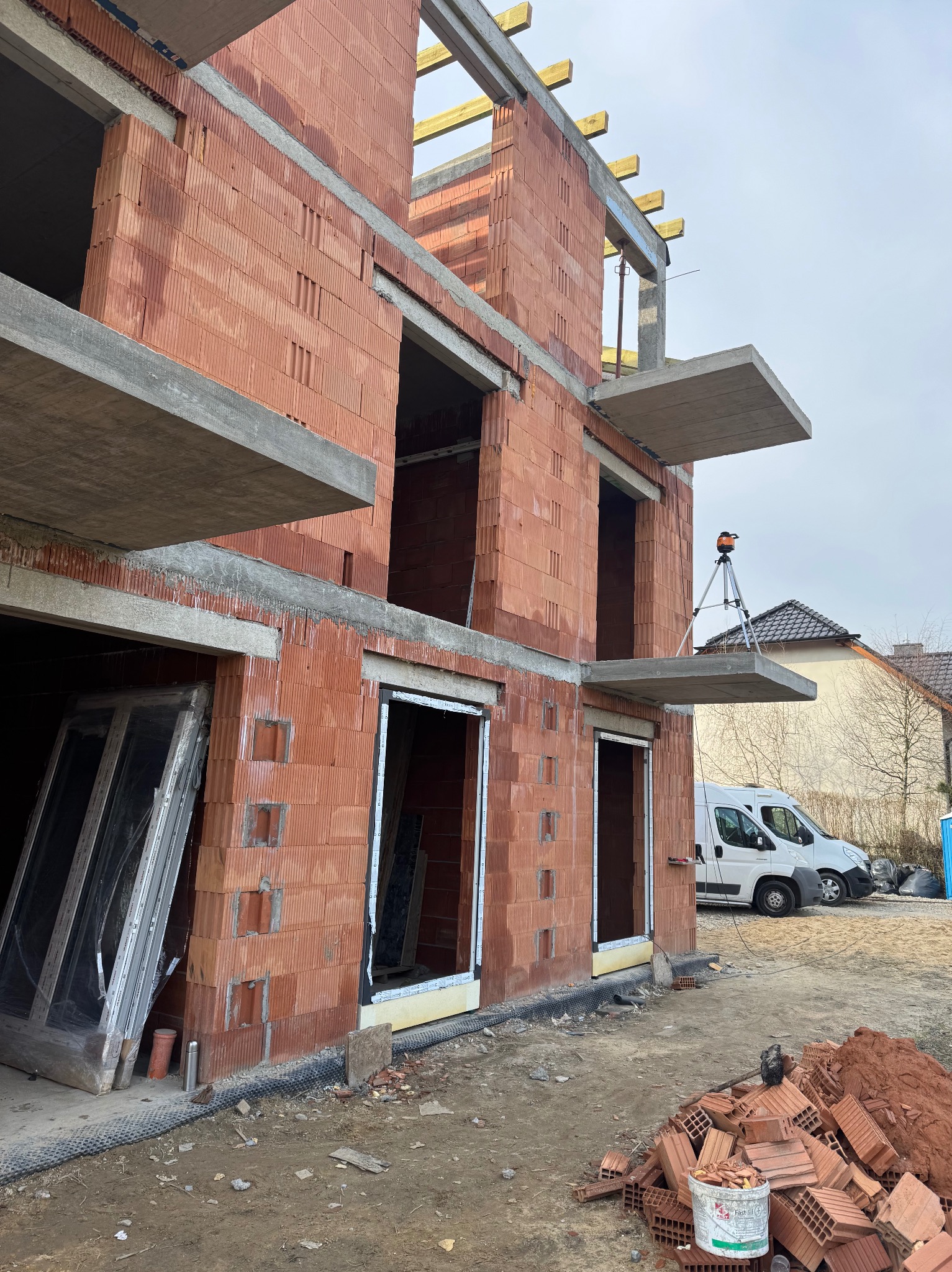 Surowy stan budynku z czerwonej cegły z oknami bez szyb, laserem na statywie i stosem cegieł. Dwa busy w tle. Widok z dołu.