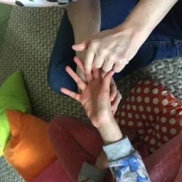 Dwie osoby siedzą na podłodze, jedna osoba masuje dłoń drugiej. Widoczne kolorowe poduszki i dziergany dywan.