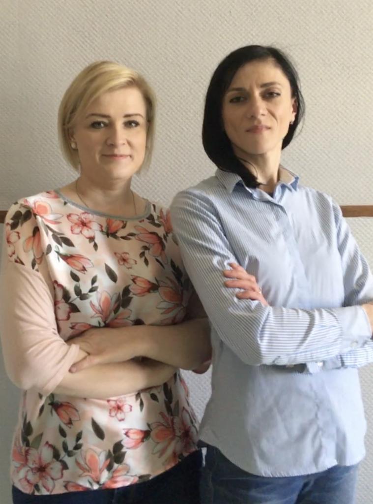 Portret dwóch kobiet z założonymi rękami na tle jasnej ściany: blondynka w bluzce w kwiaty i brunetka w błękitnej koszuli w paski.