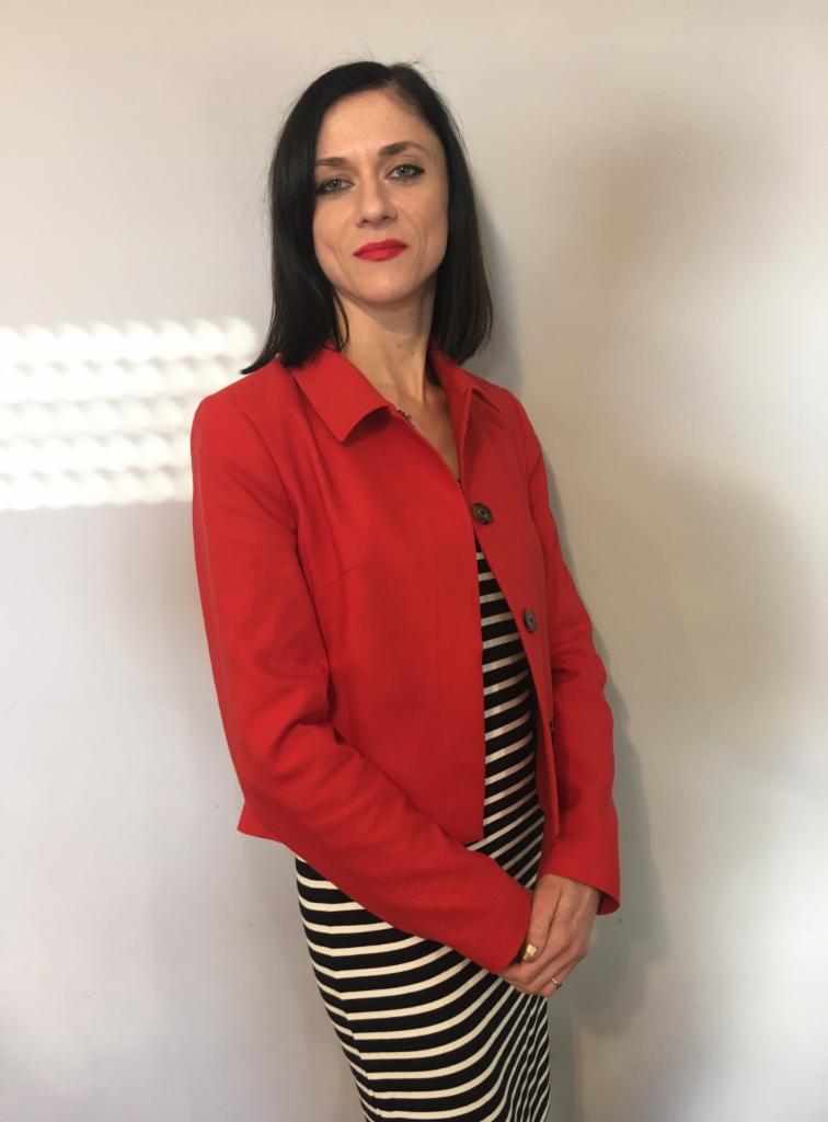 Kobieta w czerwonej marynarce i sukience w paski, stojąca na tle jasnej ściany z cieniem.