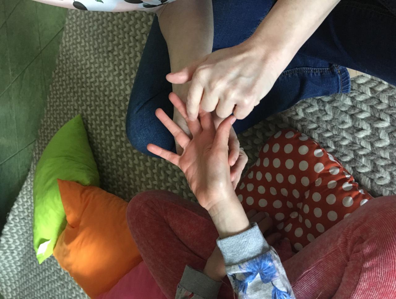 Dwie osoby siedzą na podłodze, jedna osoba masuje dłoń drugiej. Widoczne kolorowe poduszki i dziergany dywan.