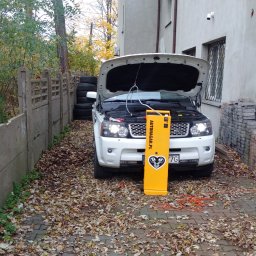 Autonagar.pl Sp.z o.o - Biały Range Rover z otwartą maską, podłączony do żółtego urządzenia do wodorowania silnika Autonagar.pl, na podwórku z liśćmi.