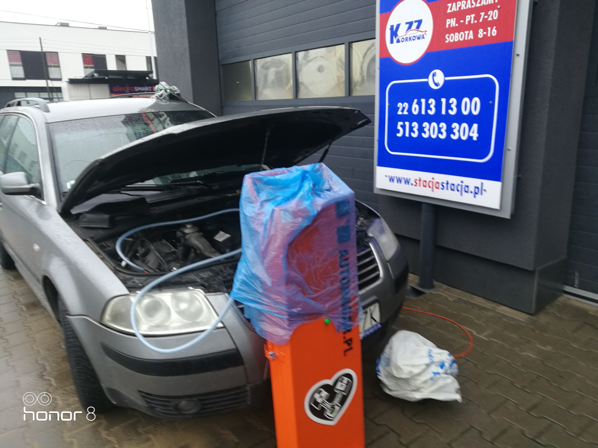 Wodorowanie silnika w samochodzie osobowym. Pod maską widoczny silnik z podłączonymi przewodami od urządzenia do wodorowania. Pomarańczowe urządzenie obok auta, zabezpieczone folią. W tle budynek...