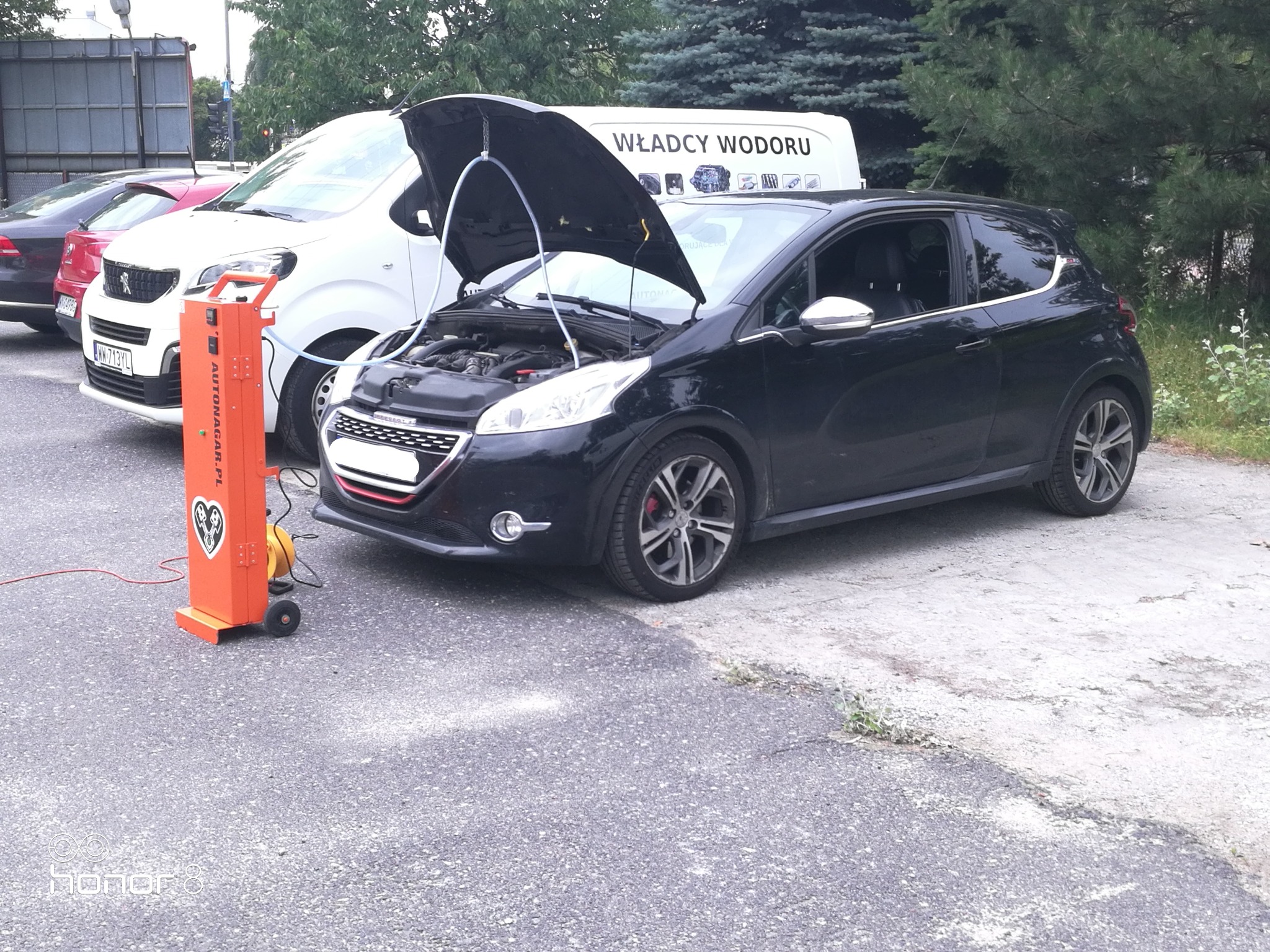 Czarny Peugeot 208 z otwartą maską, podłączony do pomarańczowego urządzenia do wodorowania silnika Autonagar.pl, na tle białego busa z napisem 'Władcy Wodoru'.