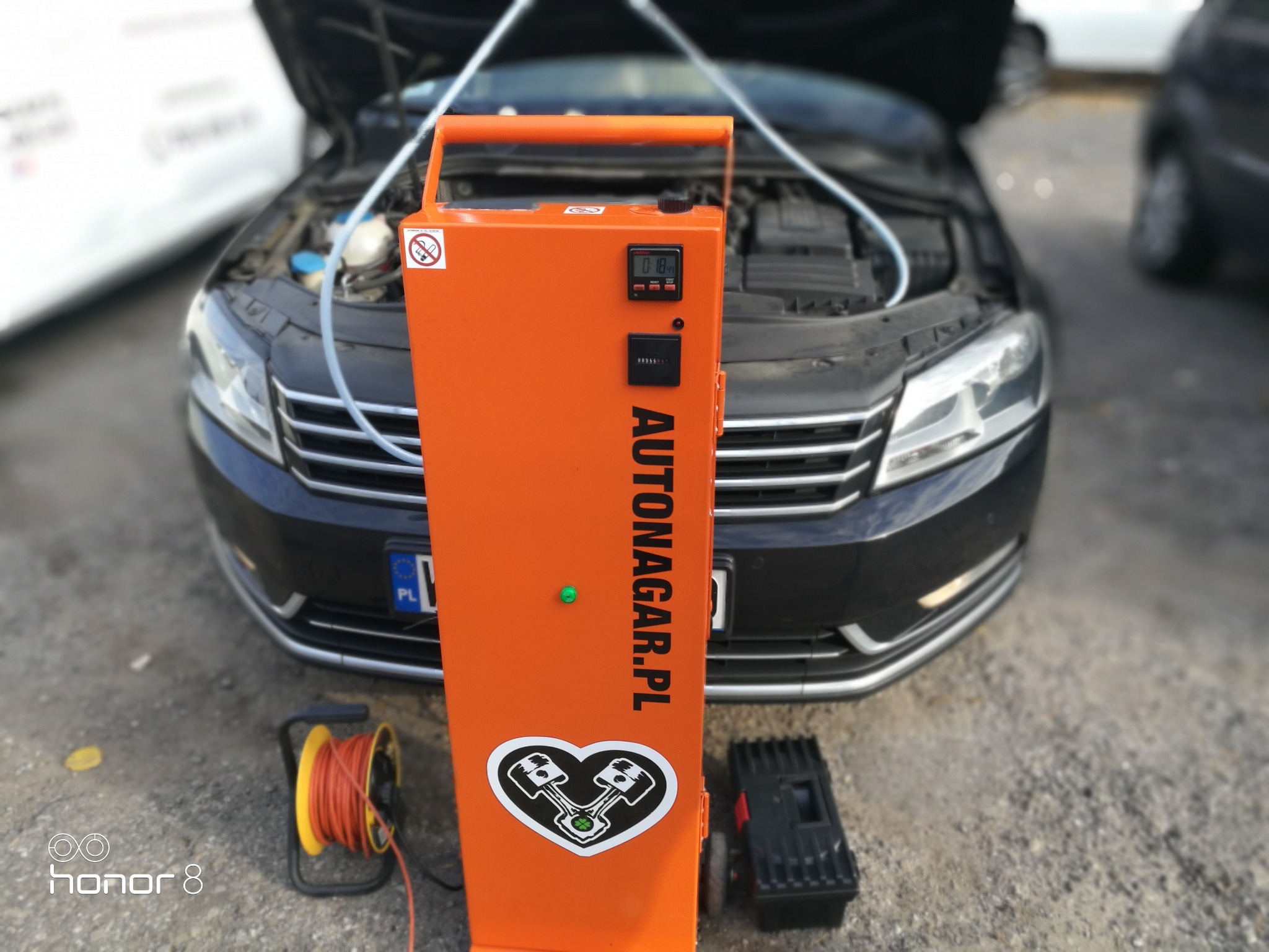 Czarny Passat TDI z otwartą maską podczas wodorowania silnika za pomocą pomarańczowego urządzenia Autonagar.pl z wyświetlaczem i naklejką z motywem silnika w sercu.
