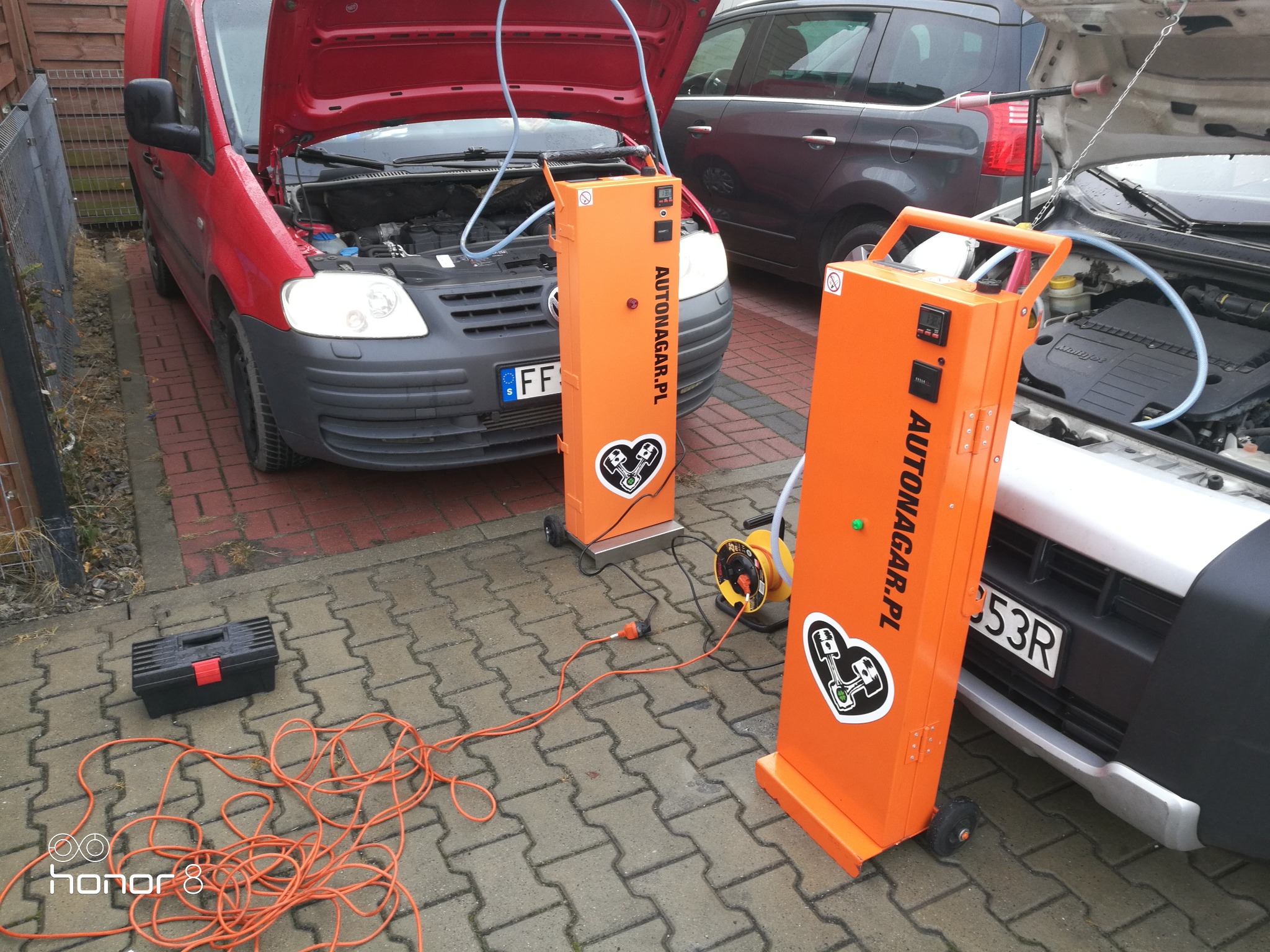 Wodorowanie silników dwóch samochodów: czerwonego i srebrnego, za pomocą dwóch pomarańczowych urządzeń Autonagar.pl z widocznymi przewodami i skrzynką narzędziową obok.
