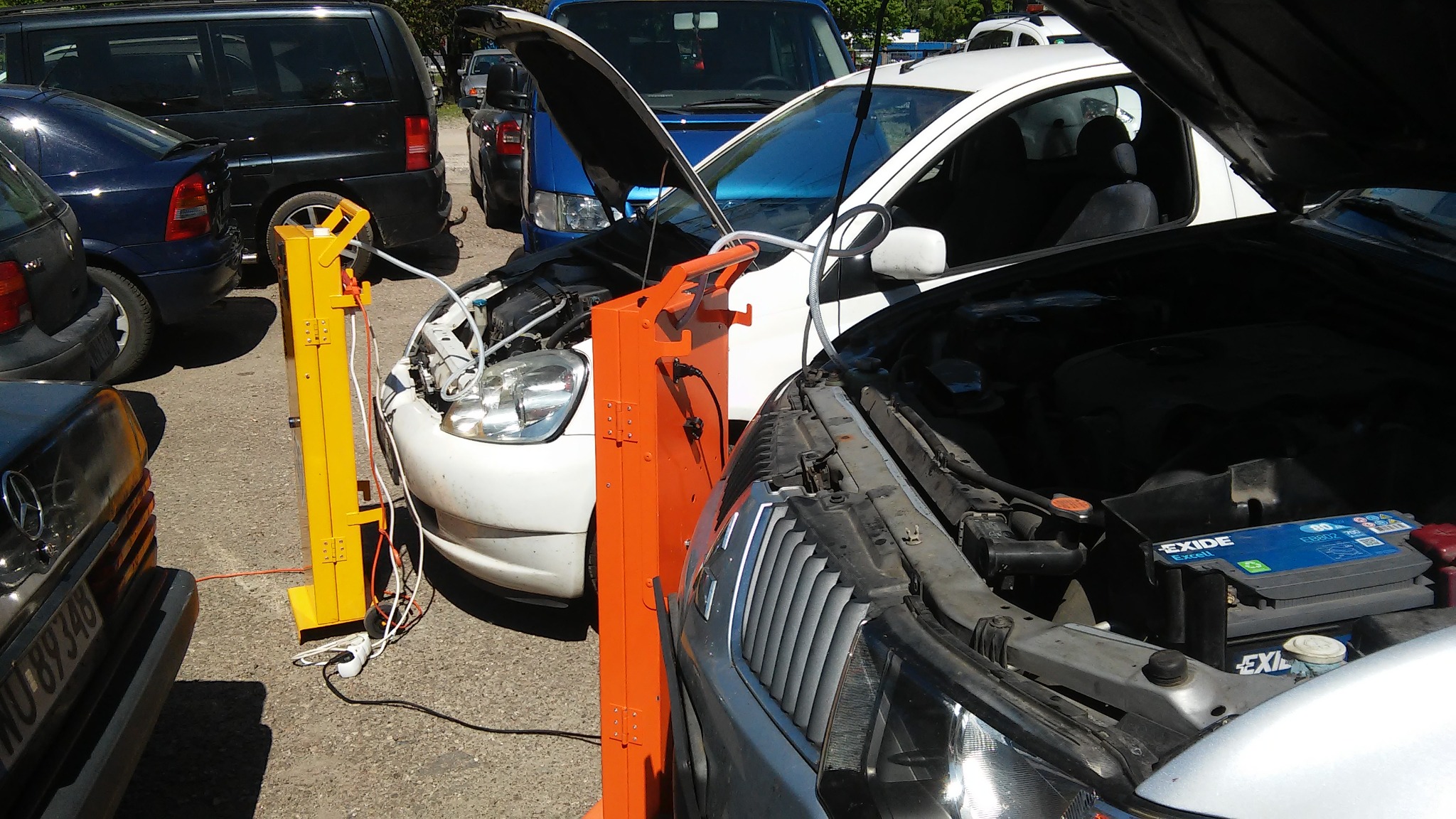 Dwa samochody z otwartymi maskami, podłączone do przenośnych urządzeń do wodorowania silnika w kolorach pomarańczowym i żółtym, na parkingu w słoneczny dzień.