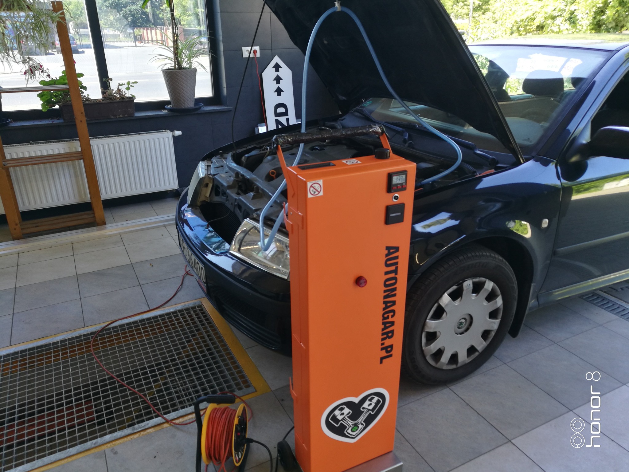 Wodorowanie silnika samochodu osobowego z użyciem pomarańczowego urządzenia Autonagar.pl z podłączonymi przewodami, widoczny otwarty przedni panel pojazdu i węże doprowadzające, w tle okno warsztatu.