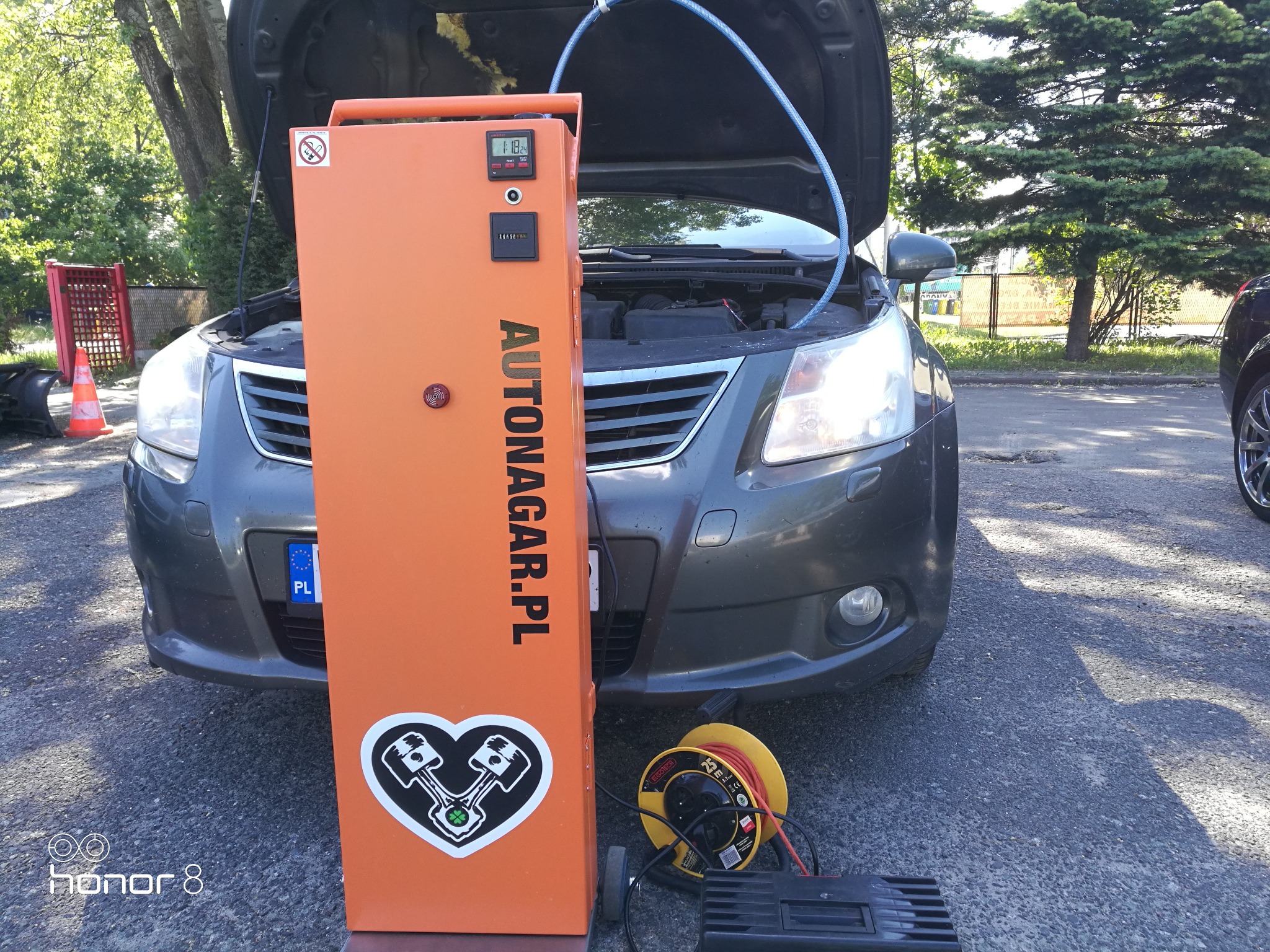 Wodorowanie silnika samochodu w Warszawie za pomocą urządzenia Autonagar.pl. Widoczny otwarty przód samochodu z podłączonym przewodem oraz pomarańczowy automat z wyświetlaczem i naklejką z logiem.