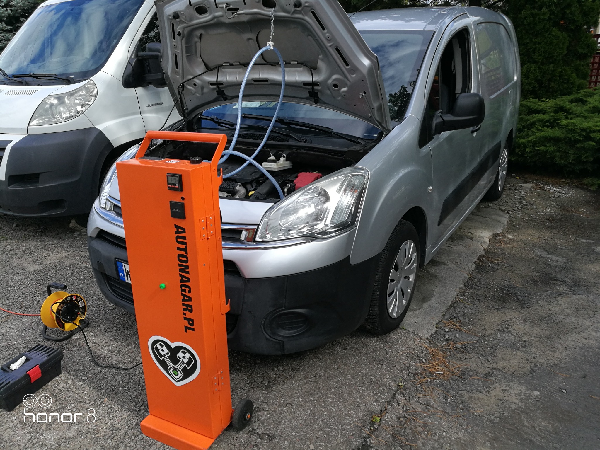 Srebrny samochód dostawczy z otwartą maską podczas procesu wodorowania silnika za pomocą pomarańczowego urządzenia Autonagar.pl; widoczny drugi samochód w tle.