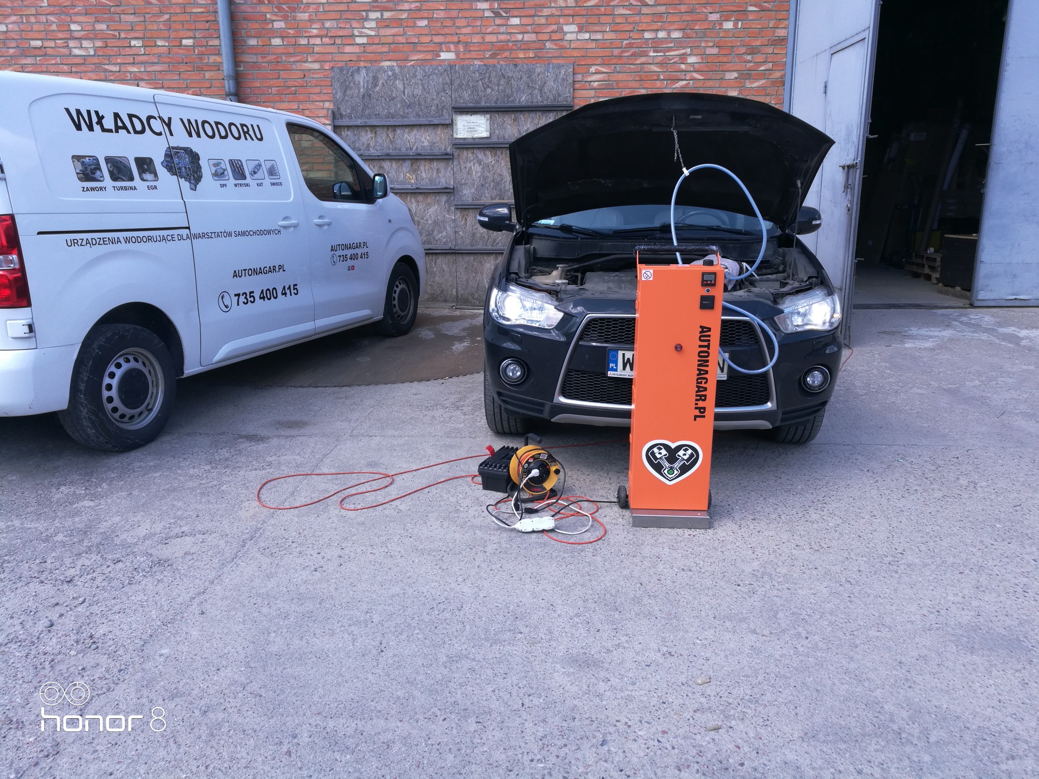 Czarny samochód z otwartą maską podłączony do pomarańczowego urządzenia do wodorowania silnika Autonagar.pl, obok biały bus z reklamą wodorowania, na betonowym placu przed budynkiem.